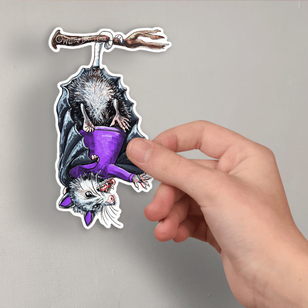 Photo of a hand holding a sticker of an illustration of an opossum dressed in a bat costume hanging upside down.