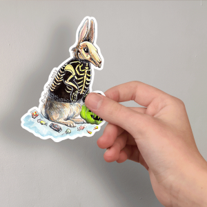 Photo of a hand holding a sticker of an illustration of a rabbit wearing a skeleton costume with a plastic Jack O'Lantern candy bucket and candy scattered around them.