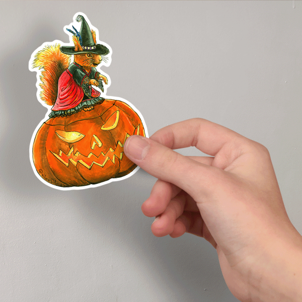 Photo of a hand holding a sticker of an illustration of a squirrel wearing a witch costume sitting on a Jack O'Lantern.