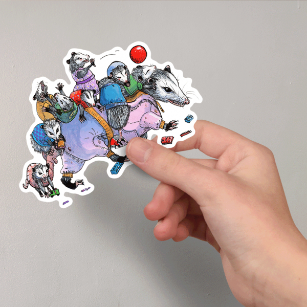 Photo of a hand holding a sticker of an illustration of an opossum carrying her many children on her back, they are all wearing cute clothes and the momma has just stepped on a toy block.