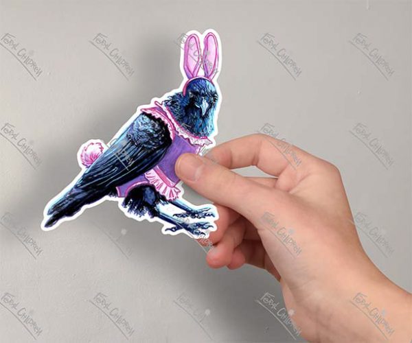 A photo of a hand holding a sticker of an illustration of a crow wearing a pink bunny costume complete with ears and tail and a ballet tutu.