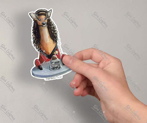 A photo of a hand holding a sticker of an illustration of a deer dressed as a character from a popular science fiction space exploration show sitting at a table in front of a chocolate sundae.