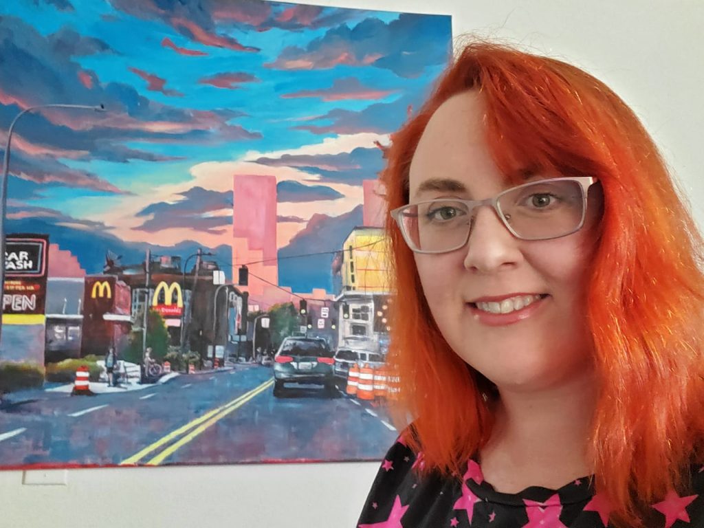 A photo of Willow D'Arcy with red hair smiling in front of her pink cityscape painting.